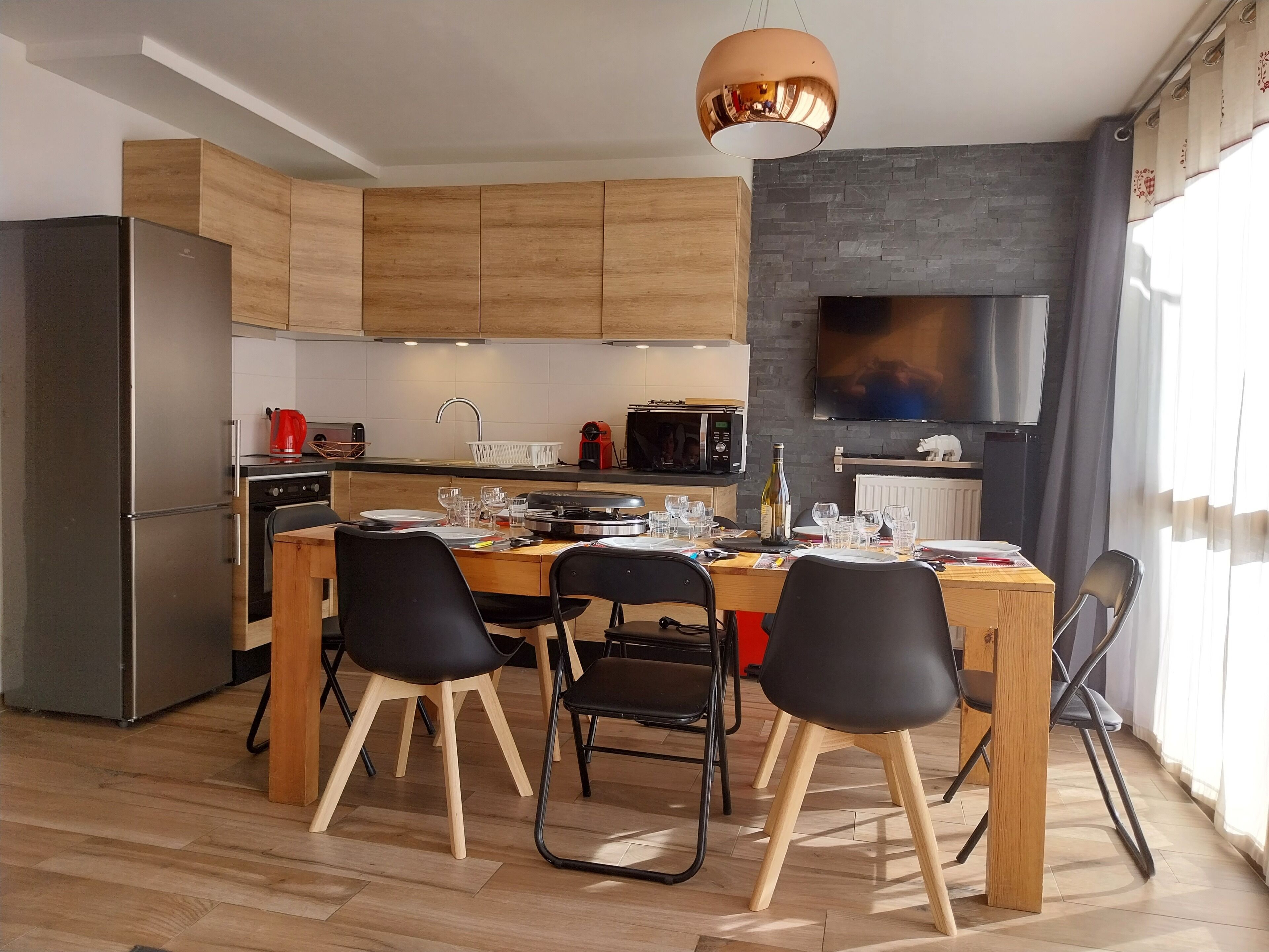 Eine moderne Küche mit Holzschränken, einem Edelstahlkühlschrank, einem Esstisch mit Stühlen und einem großen Flachbildfernseher an der Wand.