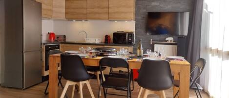 A modern kitchen with wooden cabinets, a stainless steel refrigerator, a dining table with chairs, and a large flat-screen TV mounted on the wall.