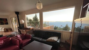 Un salon avec un canapé en cuir noir, un fauteuil rouge et une table basse. Une grande fenêtre offre une vue sur les arbres et les montagnes.
