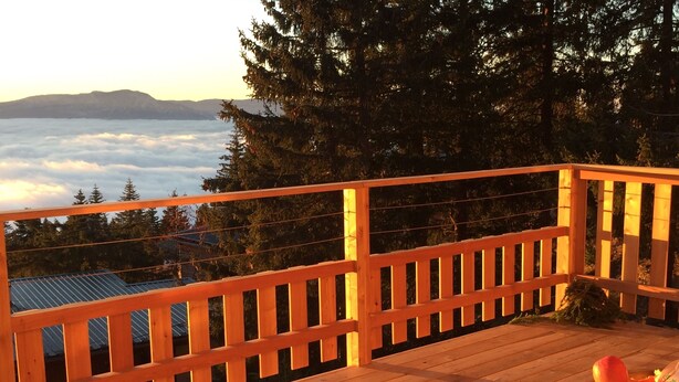 Une terrasse en bois dotée d’une balustrade, qui offre une vue sur une zone boisée et sur les nuages en contrebas.