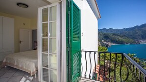 Een balkon met een bed, een groene deur en uitzicht op een kustplaatsje en bergen.