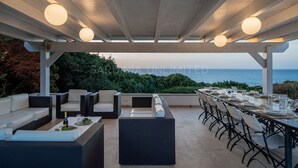 Un área de comedor al aire libre con vista al mar, asientos en porche cubierto y una pérgola con luces colgantes.