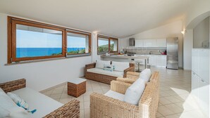 Una cocina moderna con armarios blancos y un refrigerador de acero inoxidable. En la sala de estar, hay sofás de mimbre y una mesa de comedor.