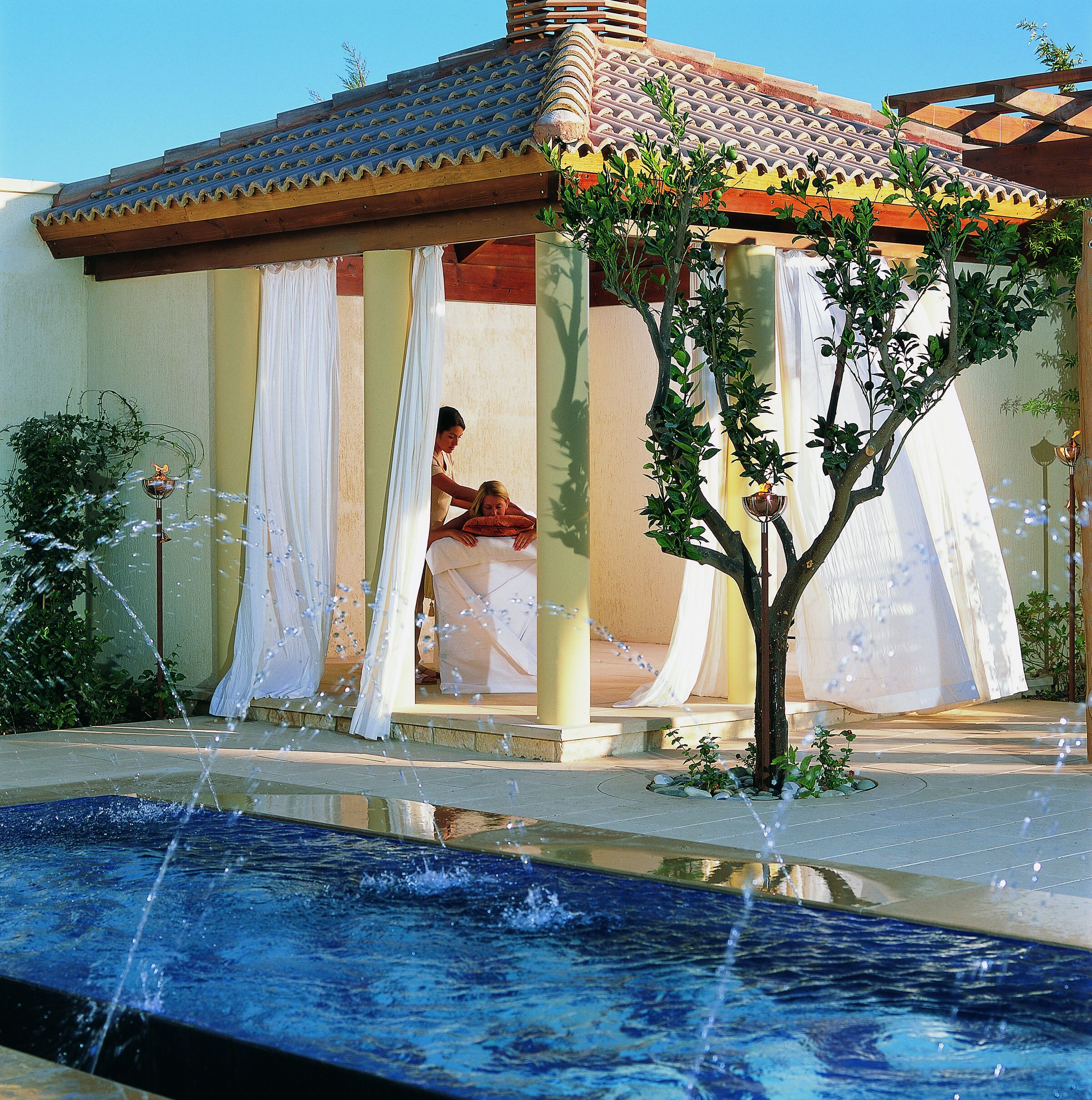 A spa area with a pool, a massage room, and a pergola with draped fabric.