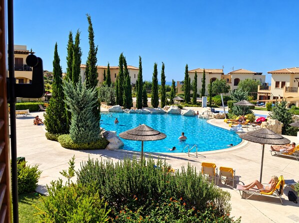 A swimming pool with lounge chairs and umbrellas, surrounded by greenery and buildings. - Theseus Village - 2-Bedroom Apartment with Pool View, Aphrodite Hills, Kouklia (Kouklia)