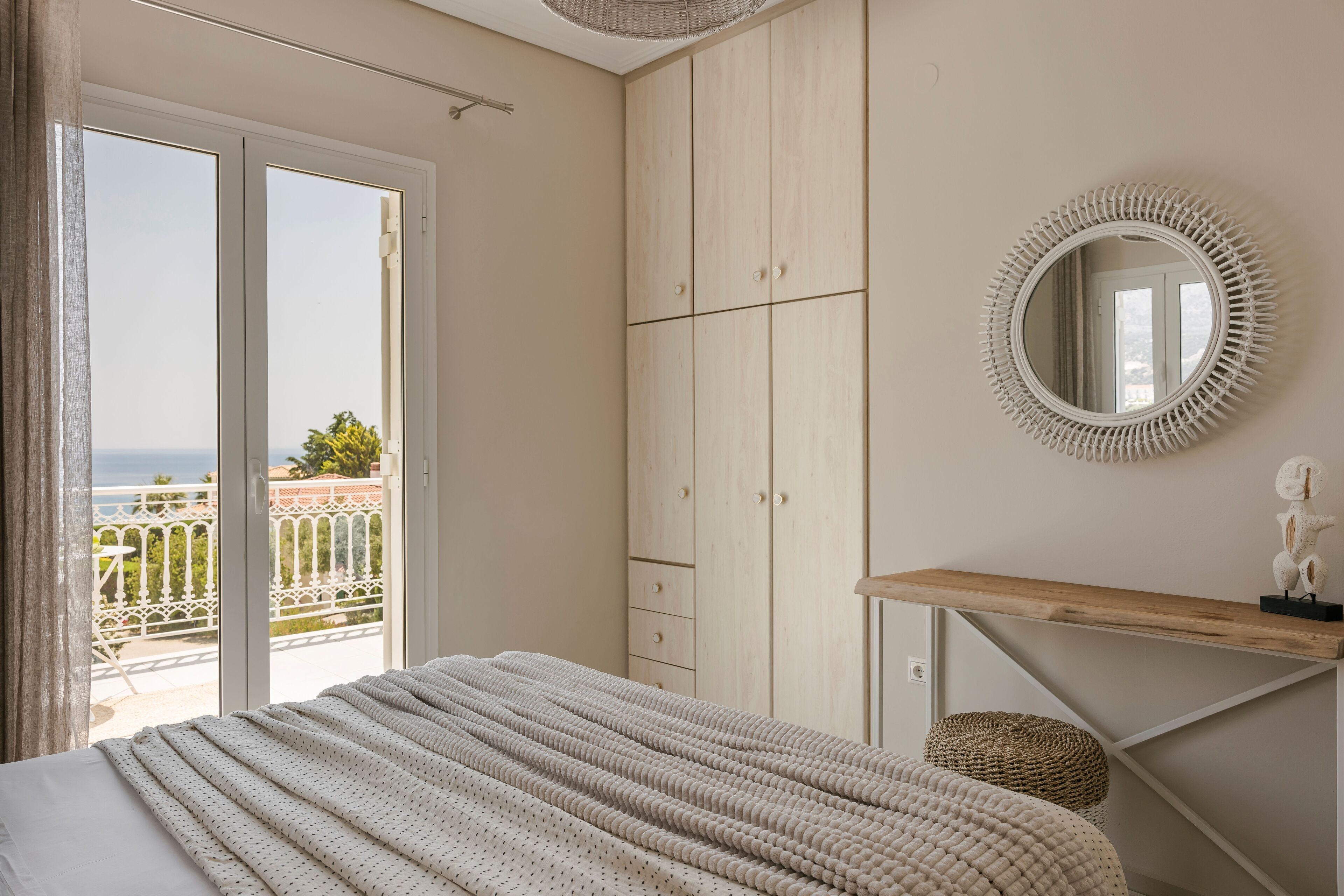 A bedroom with a bed, a wooden wardrobe, a round mirror, and a view of the outdoors.