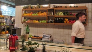 Een restaurant interieur met een krijtbordmenu, planken vol fruit en groenten, en een toonbank met drinkglazen en flessen.