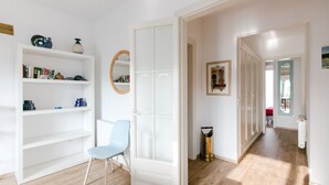 Ruime kamer met een lichtblauwe stoel, witte planken met boeken en decoratieve spullen, een ronde spiegel en een deur naar een andere kamer.