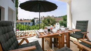 Een houten tafel met een zwart parasol, omringd door grijze gestoffeerde stoelen, op een balkon met uitzicht op gebouwen en begroening.