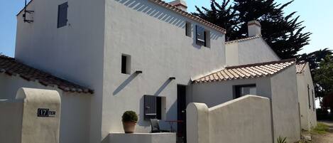 Un bâtiment à deux étages, doté d’un toit en tuiles, d’un petit jardin agrémenté d’une plante en pot et d’une porte bien visible.