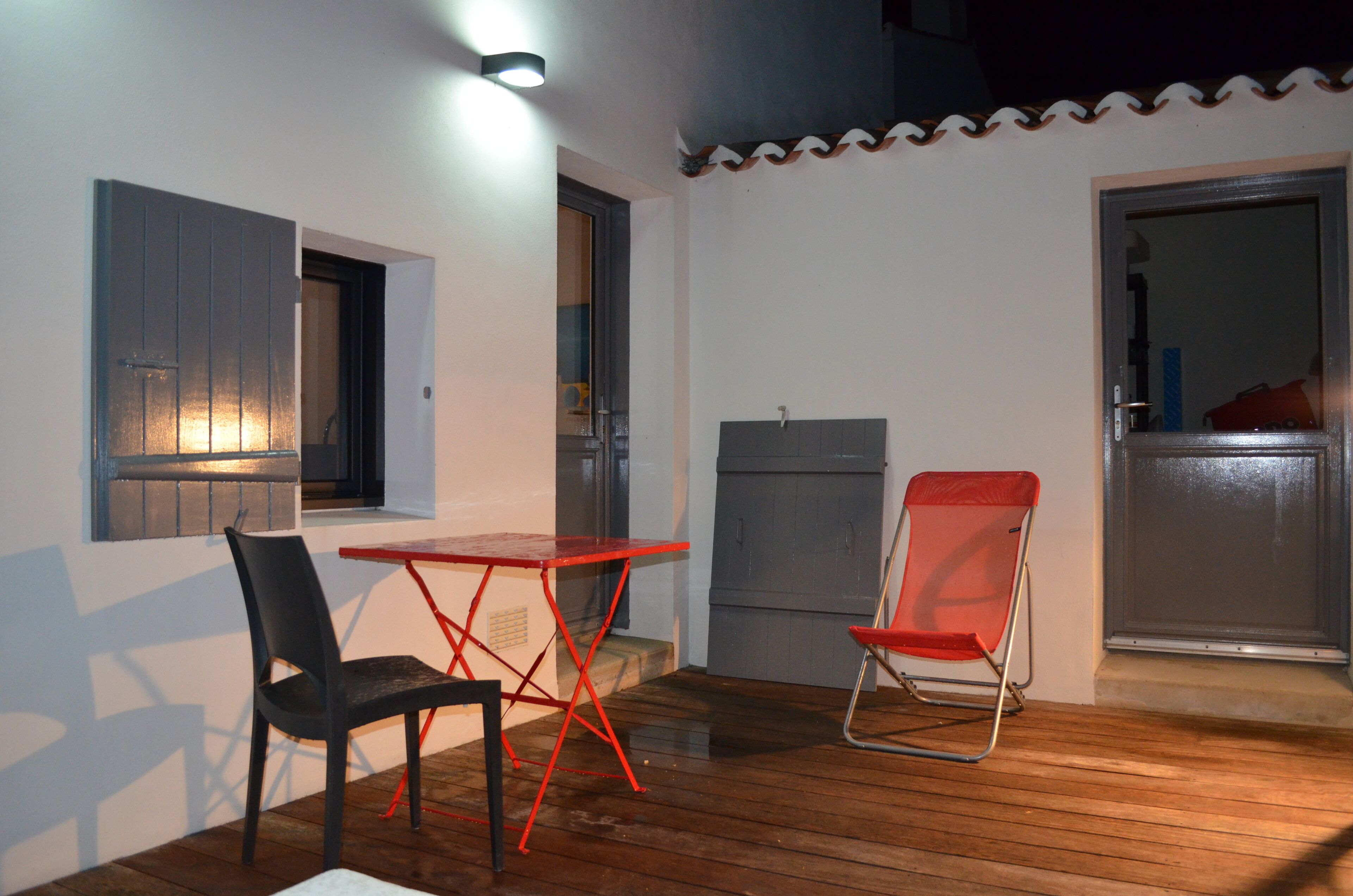 Une terrasse avec une table et deux chaises, une porte et une fenêtre avec un luminaire.