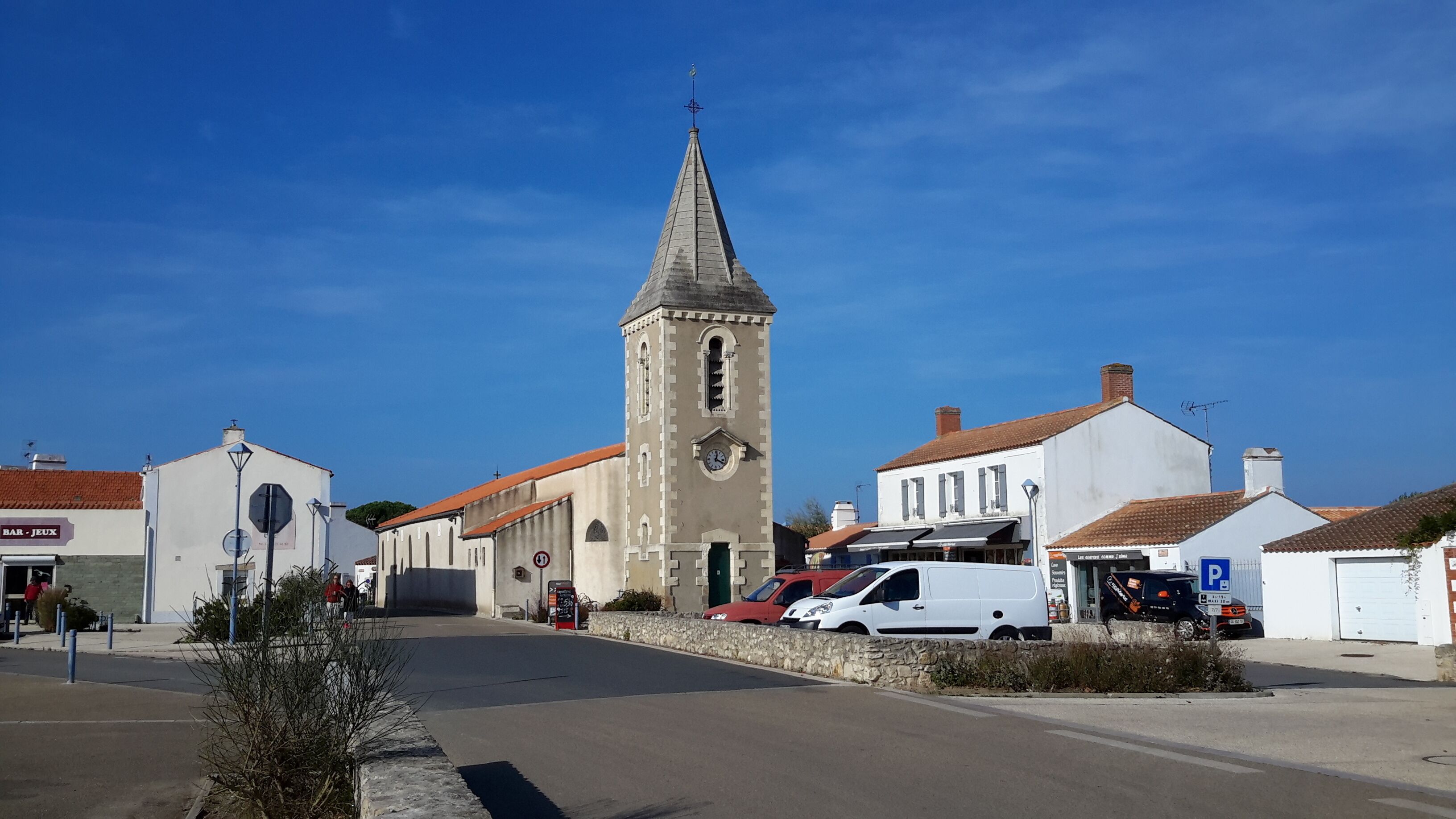 A street with a church tower, a white building with a red roof, and a parking sign.