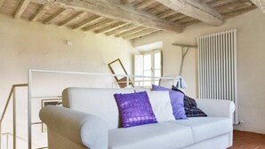 Un sofá blanco con cojines morados y azules en una habitación con vigas de madera a la vista y un radiador en la pared.