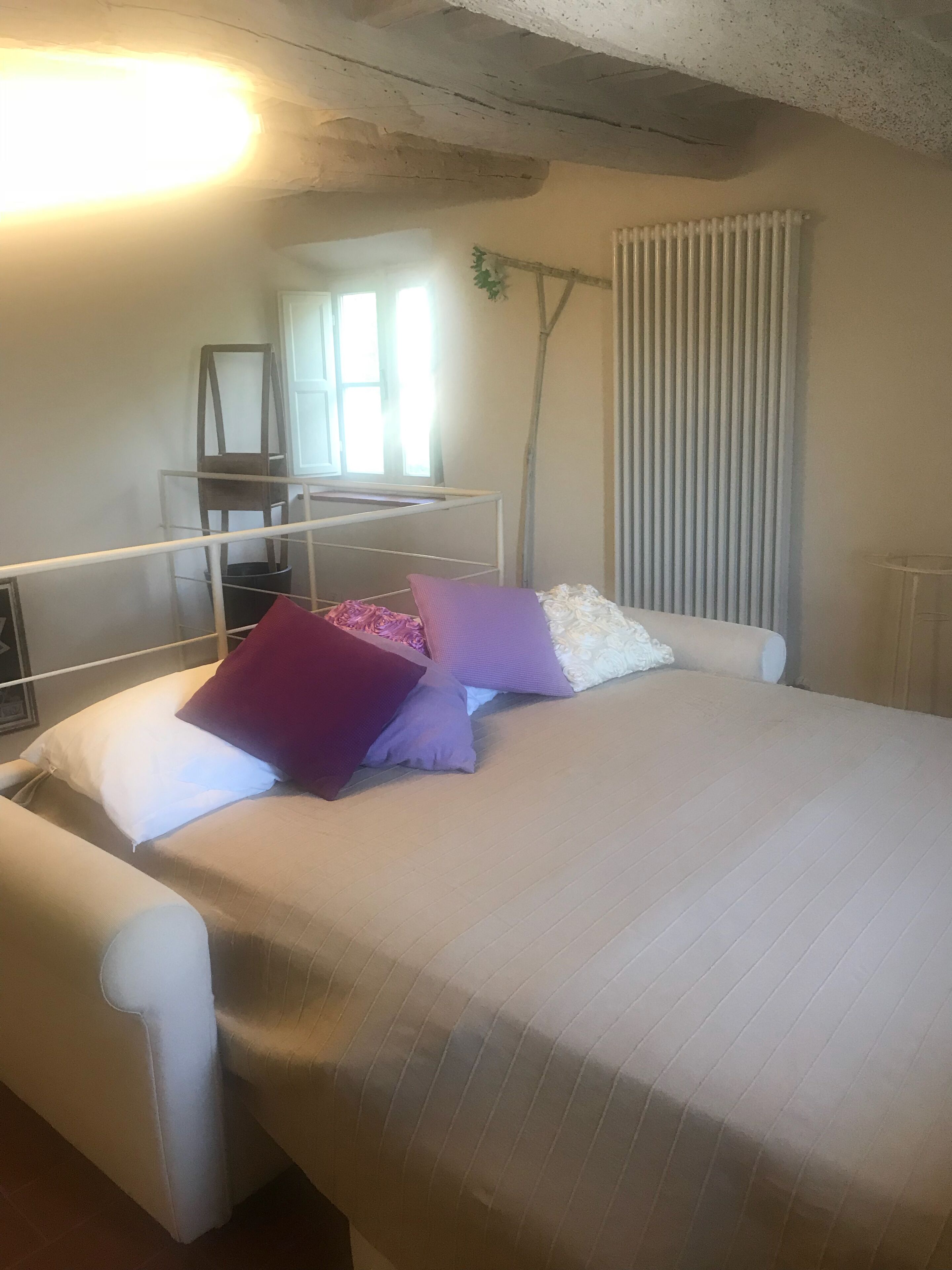 A bed with white bedding and multiple pillows, a white chair, a radiator, and a window with blinds.