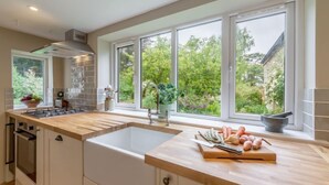A modern kitchen with a large island, a built-in oven, and a window with a view of greenery. - Tusker House, Wells-next-the-Sea, Norfolk (Wells-next-the-Sea)