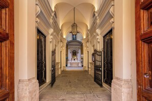 A grand hallway with ornate wooden doors and columns, leading to a central area with a statue and a chandelier.