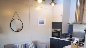 A compact living space with a sofa, armchair, and a kitchen area with modern appliances. - The Crow's Nest - A Cosy Village Cottage with Stunning Sea Views (Cawsand)