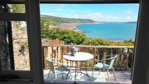 A balcony with a table and chairs, overlooking a coastal landscape with hills and the sea. - The Crow's Nest - A Cosy Village Cottage with Stunning Sea Views (Cawsand)