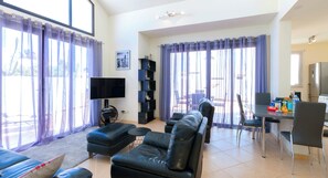 Una sala de estar moderna con televisor de pantalla plana, sofás de cuero, una zona de comedor con mesa y sillas, y ventanas amplias con cortinas.