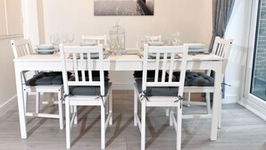 Un coin repas avec une table blanche dressée pour quatre personnes, des chaises blanches dotées de coussins gris et une vue sur la côte par la fenêtre.