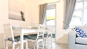 Un coin repas avec une table blanche dressée pour quatre personnes, deux chaises blanches dotées d’assises rembourrées, un canapé gris agrémenté d’un coussin à motifs et une fenêtre avec des rideaux.