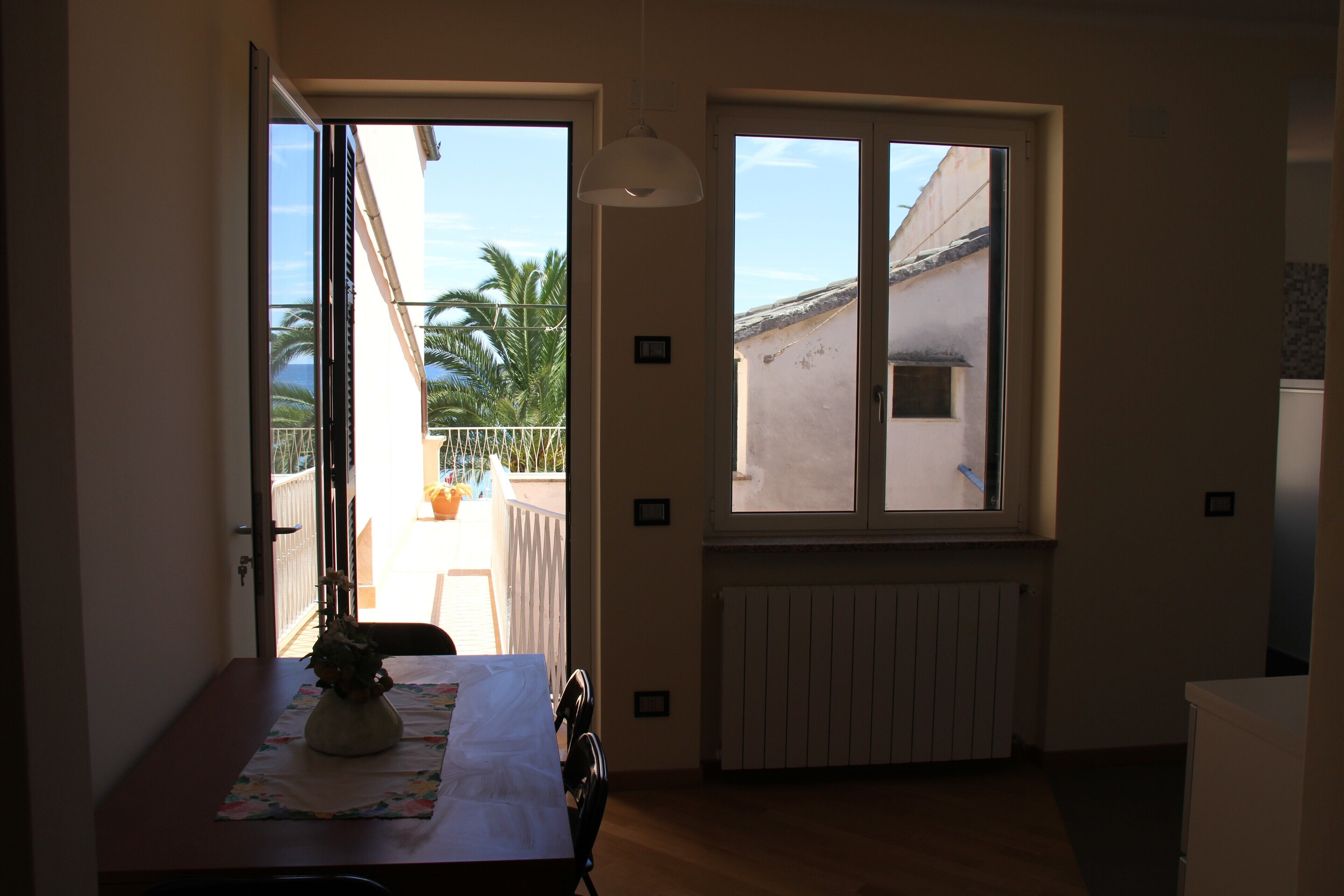 Une pièce comprenant une table à manger, des chaises, un vase de fleurs, une fenêtre donnant sur des palmiers et un immeuble, ainsi qu’un radiateur.