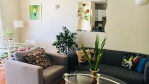 Een woonkamer met een glazen salontafel, een grijze bank, een zwarte bank en een eettafel met stoelen. Er staat een potplant op de eettafel en nog een op de salontafel.