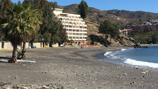 A beachfront hotel with multiple floors, a sandy beach in front, and a mountainous backdrop. - Modern villa on the beach, large private pool, BBQ, many flowers very quiet. (Almuñécar)