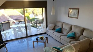 Ein Wohnzimmer mit einem beigefarbenen Sofa, einem Couchtisch und einer Glastür, die auf eine Terrasse mit weißen Stühlen und einem Tisch führt.