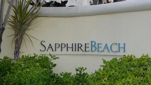 En skylt med namnet ”Sapphire Beach” på en vit vägg, omgiven av grönska.