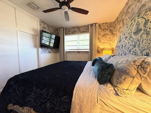Une chambre à coucher avec un lit, un ventilateur de plafond, un téléviseur fixé au mur et un papier peint à motifs.