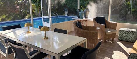 A sunlit patio with a pool, wicker furniture, and a table with lamps.