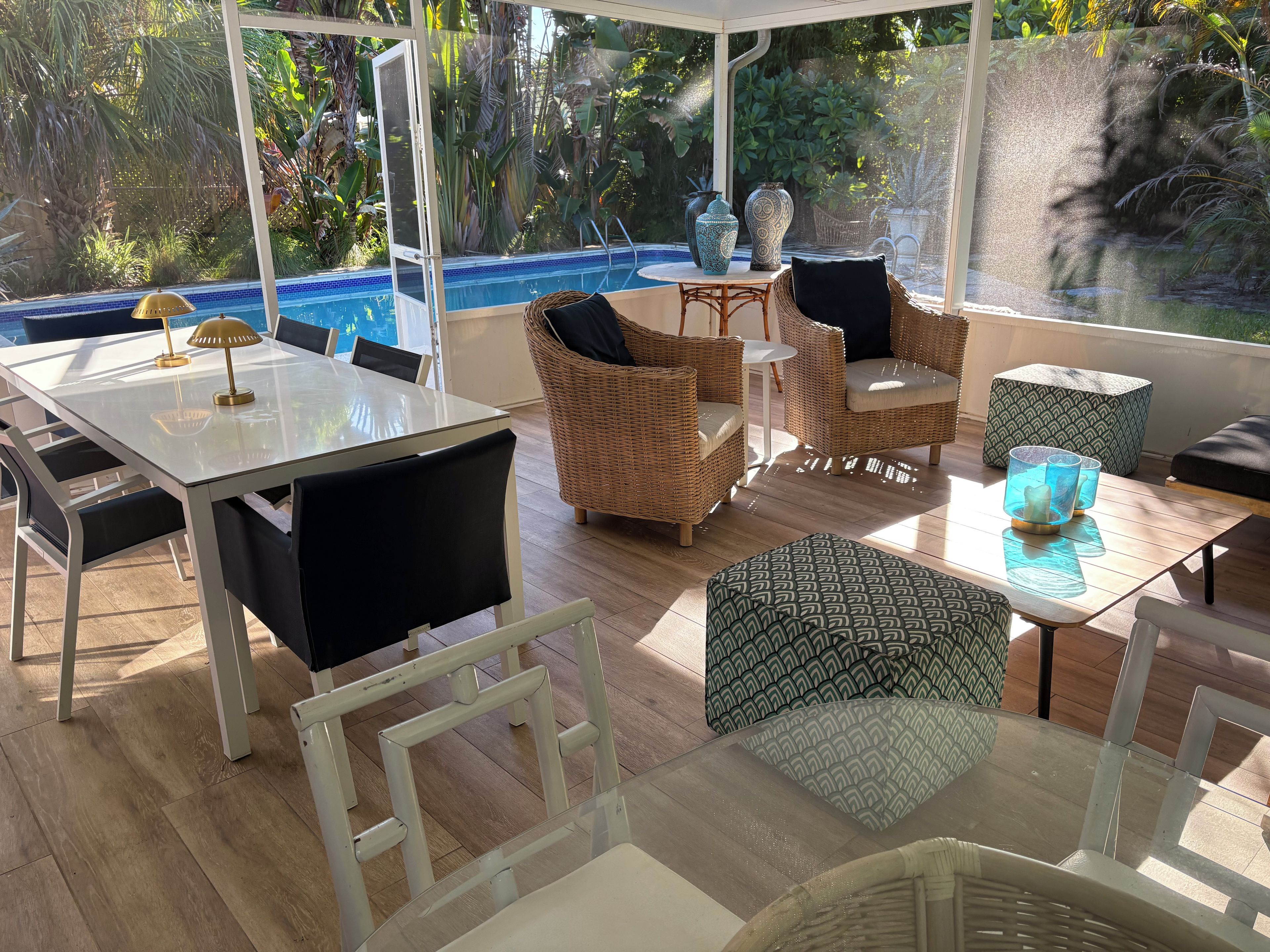 Une terrasse ensoleillée avec une table en verre, des chaises en osier et une vue sur une piscine.