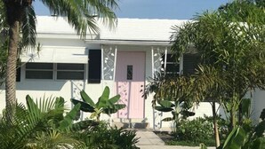 Un edificio blanco con una puerta rosa, rodeado de plantas tropicales y palmeras.