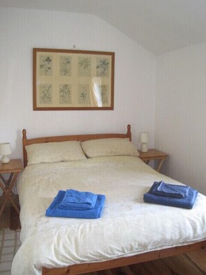 Una cama individual con estructura de madera, dos almohadas y una colcha blanca. Hay dos toallas azules dobladas sobre la cama. Encima de la cama, en la pared, cuelga un grabado botánico enmarcado.