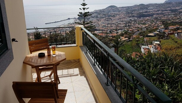 A balcony with a table and chairs, overlooking a coastal cityscape with a bridge and mountains. - Panoramic view over the city and the sea (Funchal)