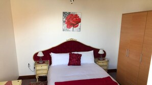 A neatly made bed with white and red bedding, a red throw blanket, and a red pillow. There is a nightstand with a lamp and a small table beside the bed. A wooden wardrobe is to the right of the bed. - Panoramic view over the city and the sea (Funchal)