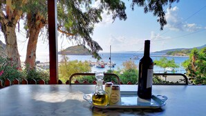 A table with a bottle of wine, two glasses, and a pepper shaker, overlooking a coastal view with boats and hills. - Shipwreck - Navagio house (Zakinthos)