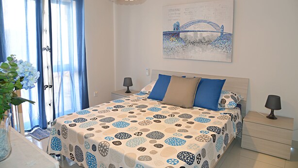 Une chambre à coucher comprenant un lit, des tables de chevet, une lampe, des rideaux, un tableau décoratif et un vase avec des fleurs.