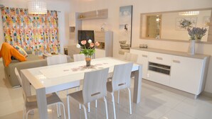 Un salon moderne avec un coin repas, comprenant une table blanche, des chaises et un canapé agrémenté d’un plaid orange.