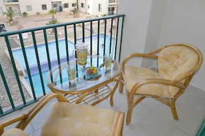 A balcony with a glass table, two wicker chairs, a fruit bowl, and a drink pitcher.