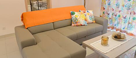 Una sala de estar moderna con un sofá seccional gris, almohadas coloridas, una manta naranja, una mesa de centro blanca con objetos decorativos y una cortina estampada con motivos florales.