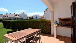 Un patio esterno con tavolo e sedie in legno, un barbecue e vista su edifici bianchi sullo sfondo.