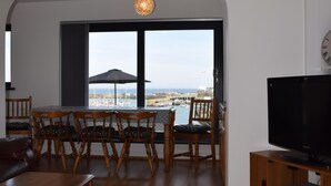 Un salon avec un plancher en bois, un coin repas avec une table et des chaises, un téléviseur et une grande fenêtre donnant sur la mer.