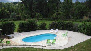 Un patio trasero con piscina, rodeado de arena y césped verde. Varias sillas de descanso y una pequeña terraza con mesa y sillas.