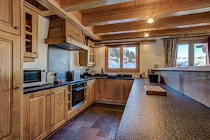 A wooden kitchen with built-in appliances, a large window with a view of a snowy landscape, and a rustic ceiling design.