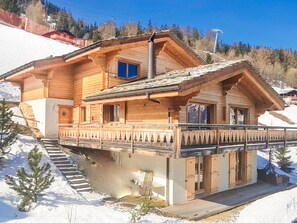A wooden chalet with a balcony, a staircase leading up to the entrance, and a view of the surrounding snowy landscape.