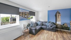 A modern living room with a grey sectional sofa, a round wooden side table, and a large window with a view of greenery. - Sparkling Clean Fabulous views of Stirling Castle  (Stirling)