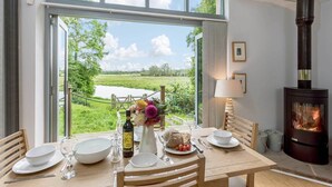 A dining area with a table set for two, a fireplace, and a view of a green landscape through large windows. - Riverside Lodge New Forest (Godshill)