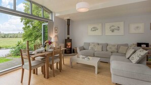 A spacious living room with a large window, a fireplace, a sofa, and a dining area. - Riverside Lodge New Forest (Godshill)